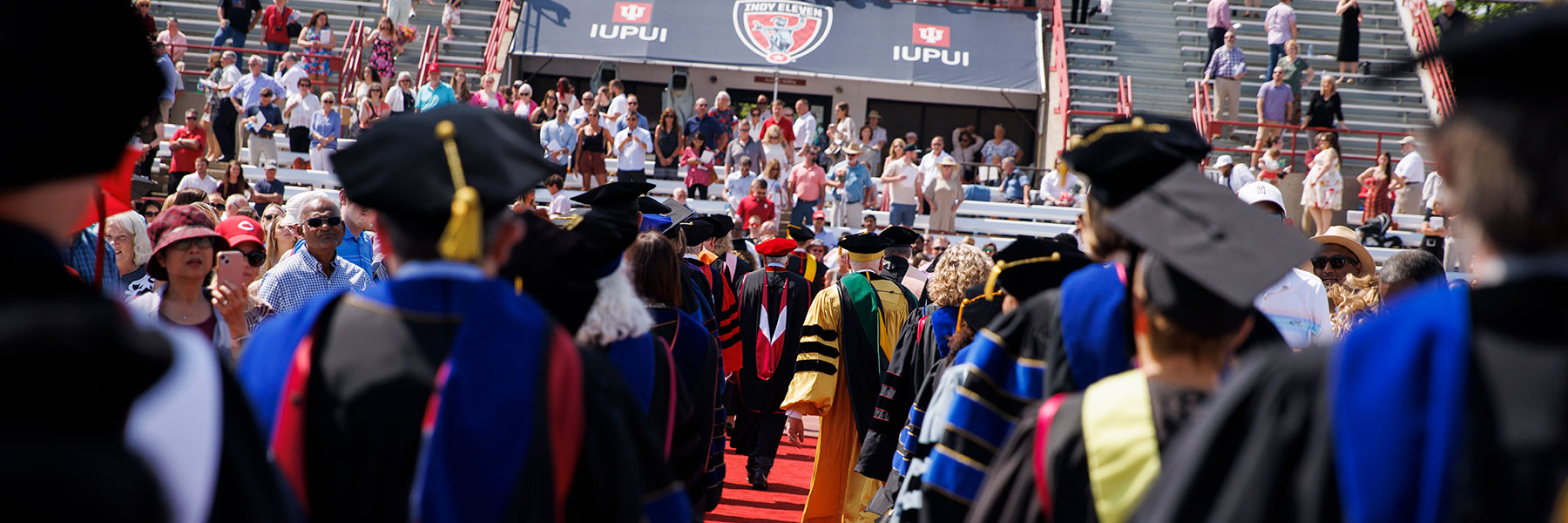 Graduating IUPUI students and faculty in graduation regalia form two lines in the pitch of Michael Carroll Stadium on campus for the 2022 commencement. Directly ahead of them is stadium seating with a crowd looking onward. Centered in the seating is a black canopy that says IUPUI and Indy Eleven